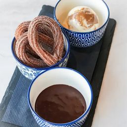 Sugared churros loops with chocolate dipping sauce and vanilla ice cream drizzled with caramel, served in blue patterned bowls on a black slate board