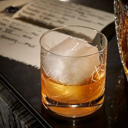 Close-up of amber whiskey served over a single large clear ice cube in an etched crystal rocks glass, set on a dark bar counter with a blurred handwritten menu in the background.
