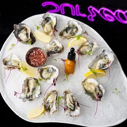 Fresh raw oysters on crushed ice served on a white platter with lemon wedges, mignonette sauce cup and a hot-sauce dropper, neon-lit seafood bar vibe.