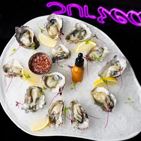 Fresh raw oysters on crushed ice served on a white platter with lemon wedges, mignonette sauce cup and a hot-sauce dropper, neon-lit seafood bar vibe.