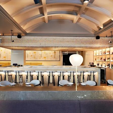 Modern upscale hotel bar interior with vaulted ceiling, illuminated marble counter, row of black bar stools, backlit liquor shelves and warm ambient lighting