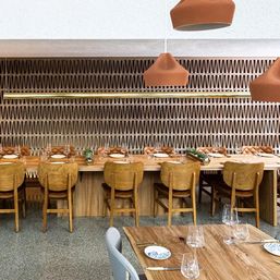 Modern restaurant interior with long wooden communal table set with plates and wine glasses, wooden chairs and tufted banquette, geometric textured wall paneling and terracotta pendant lights