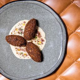 Three crispy Middle Eastern kibbeh croquettes on a creamy tahini-style sauce with herb and spice garnish, plated on a speckled gray dish set on tan quilted leather — restaurant-style appetizer.