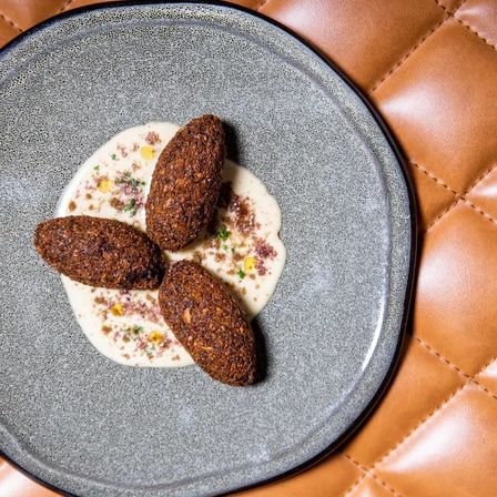 Three crispy Middle Eastern kibbeh croquettes on a creamy tahini-style sauce with herb and spice garnish, plated on a speckled gray dish set on tan quilted leather — restaurant-style appetizer.