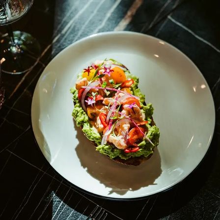 Gourmet avocado toast topped with shrimp, yellow and red cherry tomatoes, pickled red onion, microgreens and edible flowers on a white plate over a dark marble table — vibrant brunch dish.