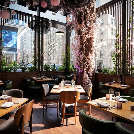 Sunlit downtown restaurant interior with wooden tables set for dining, green chairs, a cherry-blossom–wrapped column and hanging lanterns, large windows overlooking city buildings.