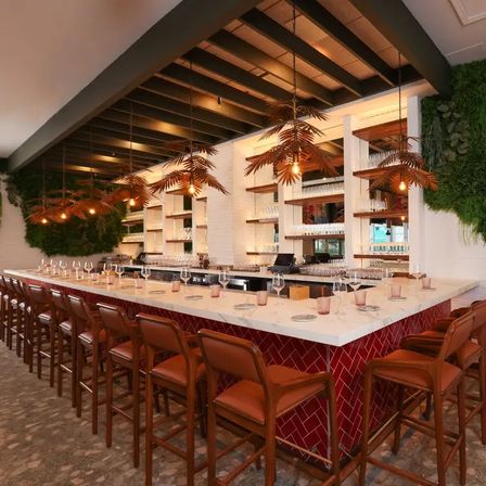Modern restaurant bar with white marble countertop and red herringbone tile front, lined with leather brown bar stools, hanging palm-leaf pendant lights, glassware-filled shelves and a living green wall.