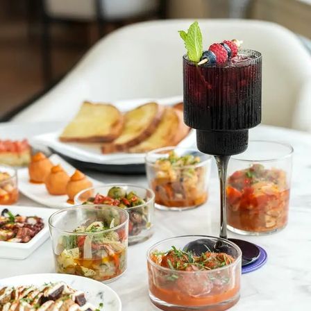 Tall black goblet cocktail garnished with mint and a berry skewer, surrounded by assorted small appetizer bowls, toasted bread and tapas-style bites on a white marble bistro table.