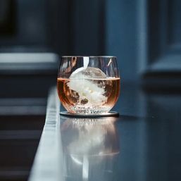 Amber craft cocktail in a round glass with a clear spherical ice orb encasing a white flower, set on a coaster on a reflective modern bar counter.