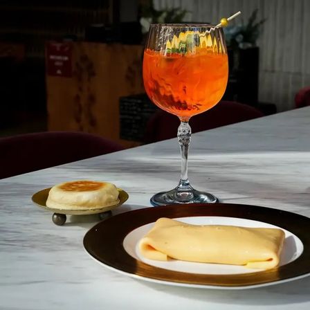 Vibrant orange aperitif in a goblet with citrus garnish on a marble bar, paired with a folded crepe on a white plate with gold rim and a small toasted pastry on a brass stand.