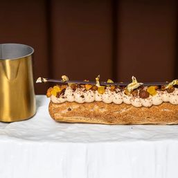 Decadent gourmet éclair on a white marble slab, topped with piped coffee cream, praline crunch, candied orange, chocolate shard and gold leaf, beside a brass pitcher.