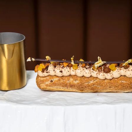 Decadent gourmet éclair on a white marble slab, topped with piped coffee cream, praline crunch, candied orange, chocolate shard and gold leaf, beside a brass pitcher.