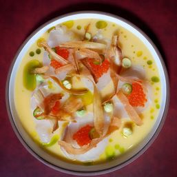 Top-down view of a gourmet scallop crudo in a citrus-yellow sauce, topped with bright salmon roe, thin crispy root chips, green herb oil droplets, jalapeño slices and dollops of avocado or wasabi cream in a white bowl on a burgundy tablecloth