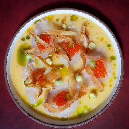 Top-down view of a gourmet scallop crudo in a citrus-yellow sauce, topped with bright salmon roe, thin crispy root chips, green herb oil droplets, jalapeño slices and dollops of avocado or wasabi cream in a white bowl on a burgundy tablecloth