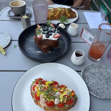 Outdoor cafe brunch: open-faced avocado toast with cherry tomatoes, microgreens, edible flowers and sliced egg on a white plate, berry-topped French toast with whipped cream on a black plate, plus omelet, coffee and a pink cocktail on a gray patio table.