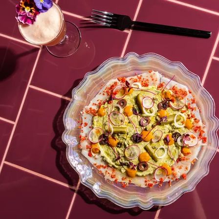 Bright avocado spread topped with radish slices, olives, yellow fruit dollops, microgreens and red crumbs on a wavy glass plate set on glossy burgundy tiles, beside a floral-garnished cocktail and black fork