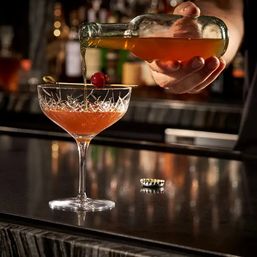 Mixologist pouring an amber cocktail from a bottle into a cut-crystal coupe glass garnished with a maraschino cherry on a dim bar counter
