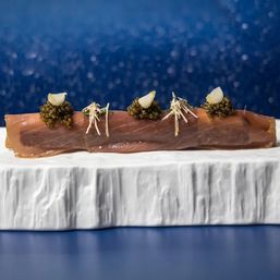 Thin slice of tuna sashimi topped with dollops of caviar, pickled garlic and shredded garnish, plated on a white marble slab against a deep-blue bokeh backdrop.