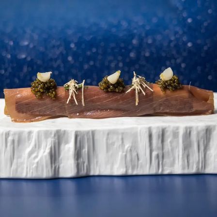 Thin slice of tuna sashimi topped with dollops of caviar, pickled garlic and shredded garnish, plated on a white marble slab against a deep-blue bokeh backdrop.