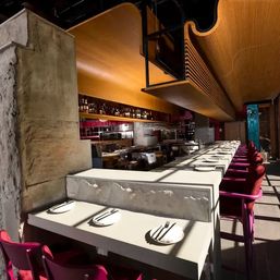Sunlit modern restaurant bar interior with long white counter, magenta bar stools, sculpted wooden ceiling, textured stone wall and set place settings.