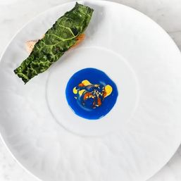 Fine-dining appetizer on a white plate: crispy kale leaf over crostini with a vivid blue sauce pool, yellow purée accents and crunchy garnish — modern plated dish.
