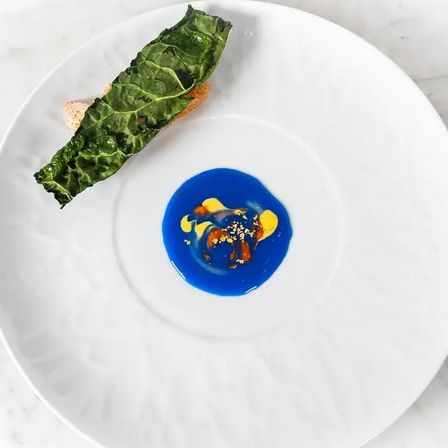 Fine-dining appetizer on a white plate: crispy kale leaf over crostini with a vivid blue sauce pool, yellow purée accents and crunchy garnish — modern plated dish.