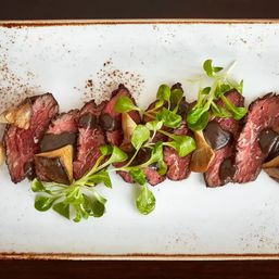 Sliced medium-rare steak drizzled with dark demi‑glace, topped with sautéed mushrooms and fresh microgreens on a rustic white rectangular plate — gourmet restaurant plating.
