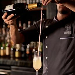 Bartender theatrically pouring sparkling wine into a champagne flute — a long ribbon of liquid suspended mid-air above foamy bubbles at an upscale cocktail bar.