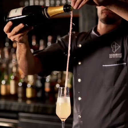 Bartender theatrically pouring sparkling wine into a champagne flute — a long ribbon of liquid suspended mid-air above foamy bubbles at an upscale cocktail bar.
