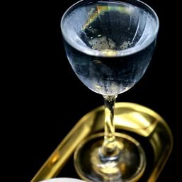 Close-up of a stemmed glass with bubbling clear liquid, perched on a shiny gold tray against a black background.
