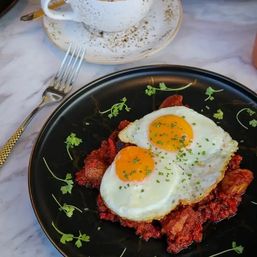 Brunch plate: two sunny-side-up eggs sprinkled with chives atop a crispy red hash, microgreen garnish on a black plate with a gold-handled fork and speckled coffee cup on a marble table.