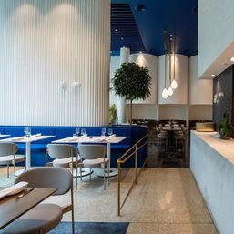 Modern upscale restaurant interior with cobalt-blue banquette seating, white ribbed wall, terrazzo floor, round tables set with glassware, brass handrail, hanging globe pendant lights and indoor tree.