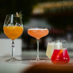 Three colorful craft cocktails on a bar counter: an orange spritz in a tall stemmed glass with herb garnish, a frothy pink coupe, and bright red and pale-yellow lowball drinks on a marble countertop.