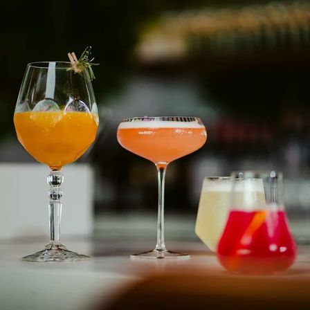 Three colorful craft cocktails on a bar counter: an orange spritz in a tall stemmed glass with herb garnish, a frothy pink coupe, and bright red and pale-yellow lowball drinks on a marble countertop.