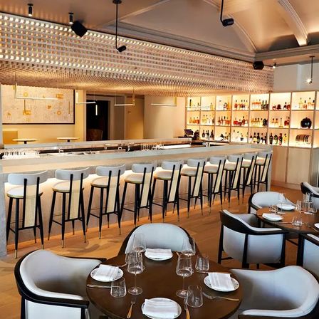 Contemporary upscale restaurant interior featuring a long marble bar with a row of white cushioned barstools, backlit liquor shelves, round dining tables set with glassware, and warm ambient lighting.