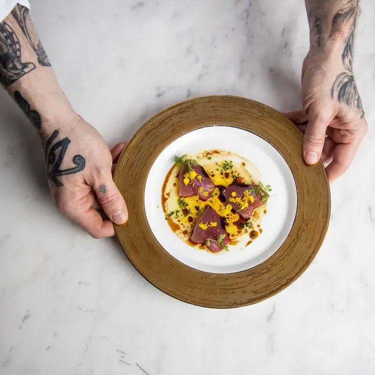 Top-down view of a fine-dining plate of seared tuna with yellow sauce, microgreens and oil drizzle on a white plate with a bronze rim, held by tattooed hands over a marble tabletop.