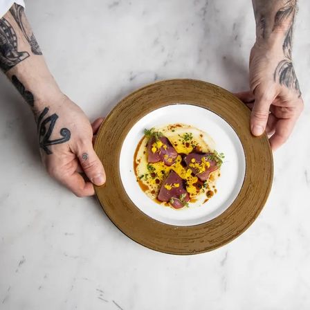 Top-down view of a fine-dining plate of seared tuna with yellow sauce, microgreens and oil drizzle on a white plate with a bronze rim, held by tattooed hands over a marble tabletop.