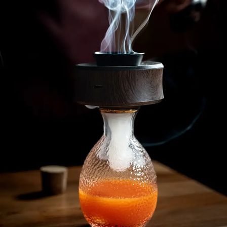 Smoked orange craft cocktail in a textured glass bulb topped by a wooden smoke infuser, wisps of aromatic smoke rising.