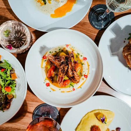 Top-down restaurant table: gourmet plate of crispy prawns on creamy purée with cherry tomatoes, surrounded by salad, omelette and wine glasses on white dishes over a wooden table.