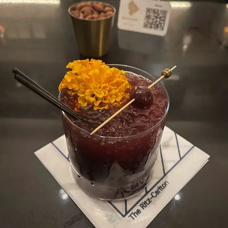 Dark red crushed-ice cocktail in a short rocks glass with two black straws, topped with a bright orange marigold and a skewered cherry, sitting on a hotel bar napkin on a dark counter.