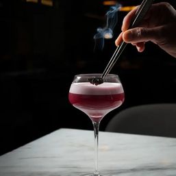 Smoke-kissed pink craft cocktail with frothy foam in a coupe glass, smoked berry garnish being placed with metal tongs on a marble bar top