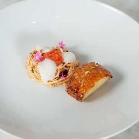 Fine-dining plate with a small nest of seafood pasta topped with foam, salmon roe and pink edible flowers, paired with a glazed, scored seared scallop on a white plate