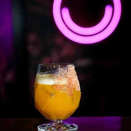 Neon-lit bar scene with a yellow-orange cocktail in a stemmed glass, colorful spiced-salt rim, and a glowing pink circular neon sign in the blurred background.