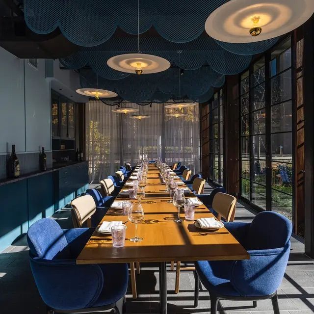 Sunlit modern restaurant dining room with a long communal wooden table set with wine glasses and napkins, blue upholstered chairs, floor-to-ceiling windows and scalloped ceiling panels with hanging pendant lights.