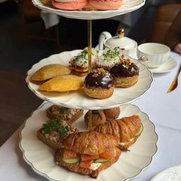 Three-tier afternoon tea stand with smoked-salmon croissant sandwiches, cucumber, savory tarts, chocolate-glazed cream puffs, mini madeleines and pink macarons beside a teapot and teacup on a white tablecloth.