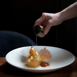 Hand pouring sauce over a gold-leaf poached pear on a white plate, garnished with caramel and a delicate sugar lattice — elegant gourmet dessert.