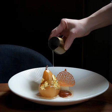 Hand pouring sauce over a gold-leaf poached pear on a white plate, garnished with caramel and a delicate sugar lattice — elegant gourmet dessert.