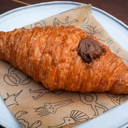 Golden, buttery chocolate croissant with a glossy chocolate dollop, resting on printed kraft paper on a white plate — flaky breakfast pastry for cafe or bakery.