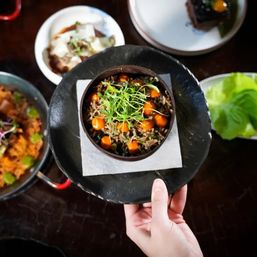 Overhead view of a gourmet Asian-style rice bowl topped with microgreens and bright orange sauce pearls, served in a dark bowl on a black plate held by hand, surrounded by small dishes on a wooden restaurant table.