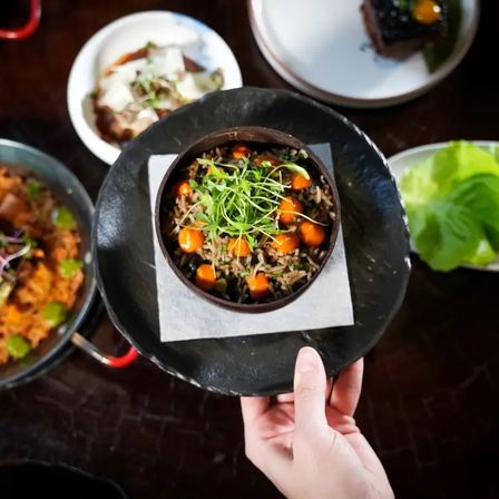 Overhead view of a gourmet Asian-style rice bowl topped with microgreens and bright orange sauce pearls, served in a dark bowl on a black plate held by hand, surrounded by small dishes on a wooden restaurant table.